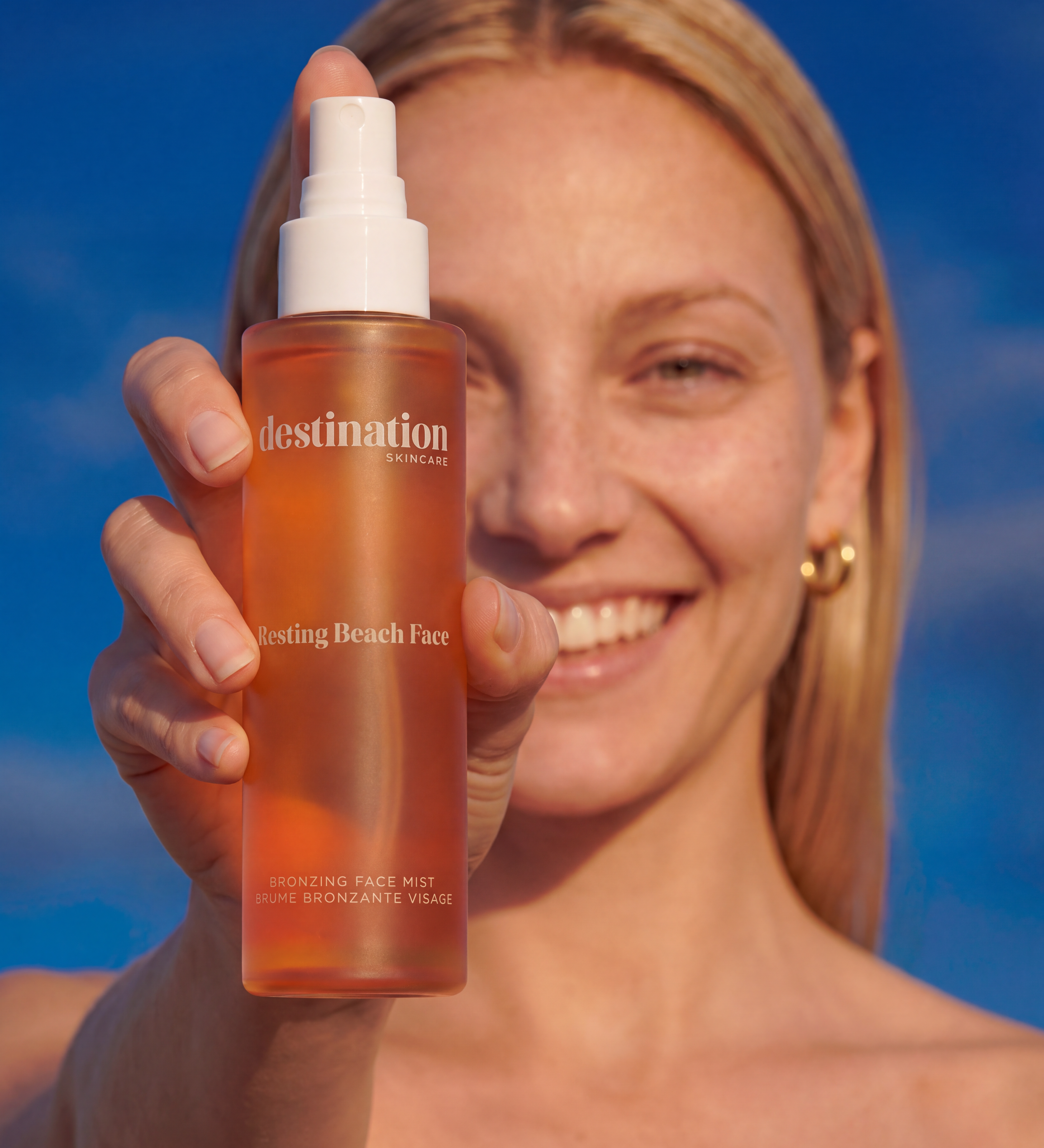 Generated_with_Kive.ai_-_Modify_the_pose_so_the_woman_extends_one_hand_and_arm_toward_the_camera__holding_the_bronzing_face_mist_prominently_in_the_extreme_foreground_between_the_came.png